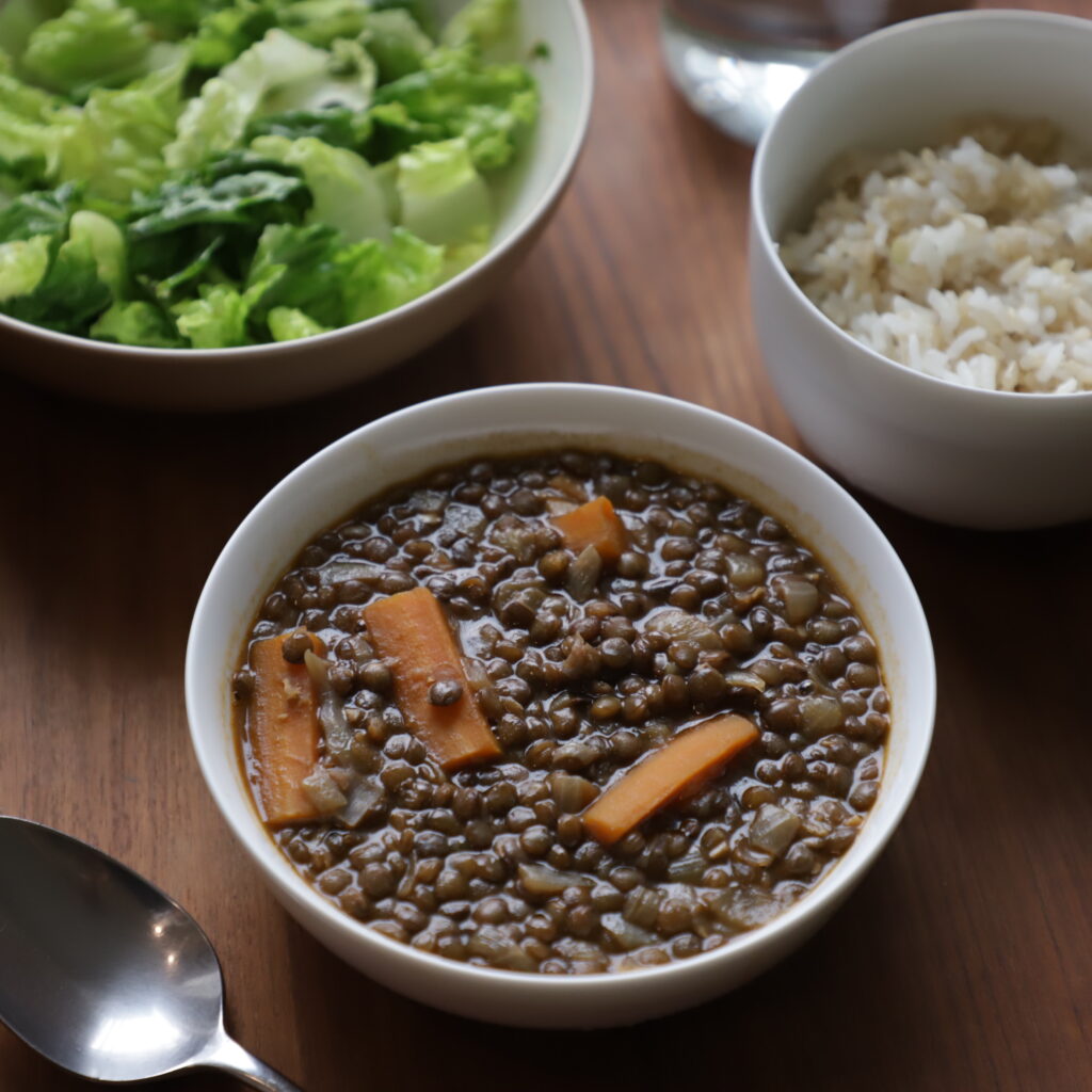 another bowl of reddish-brown stewed French green lentils next to a bowl of bright green lettuce and a bowl of brown rice, with a spoon in the lower corner