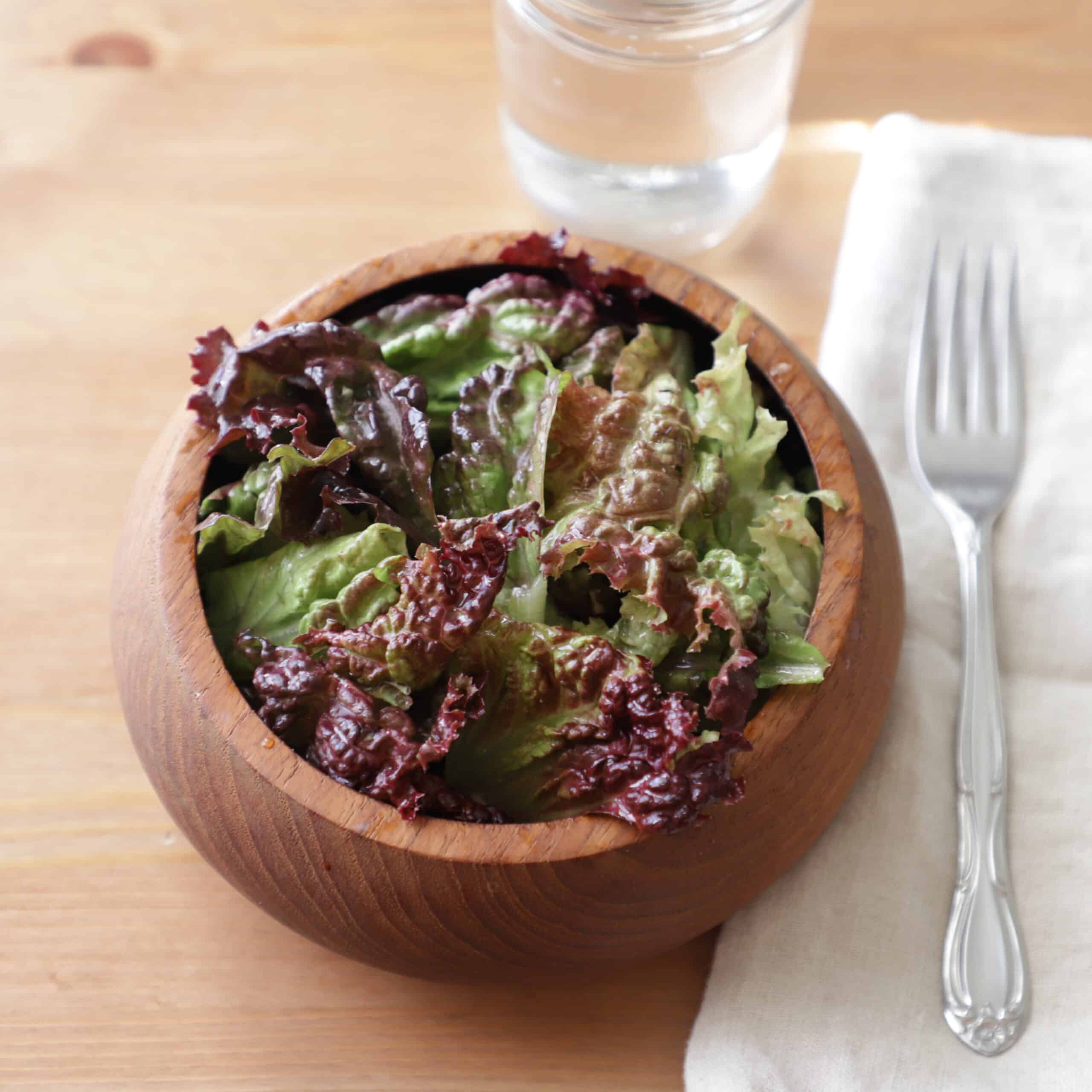 Rounded dark wooden bowl containing red leaf lettuce, next to a fork, napkin, glass of water and on a wooden surface.
