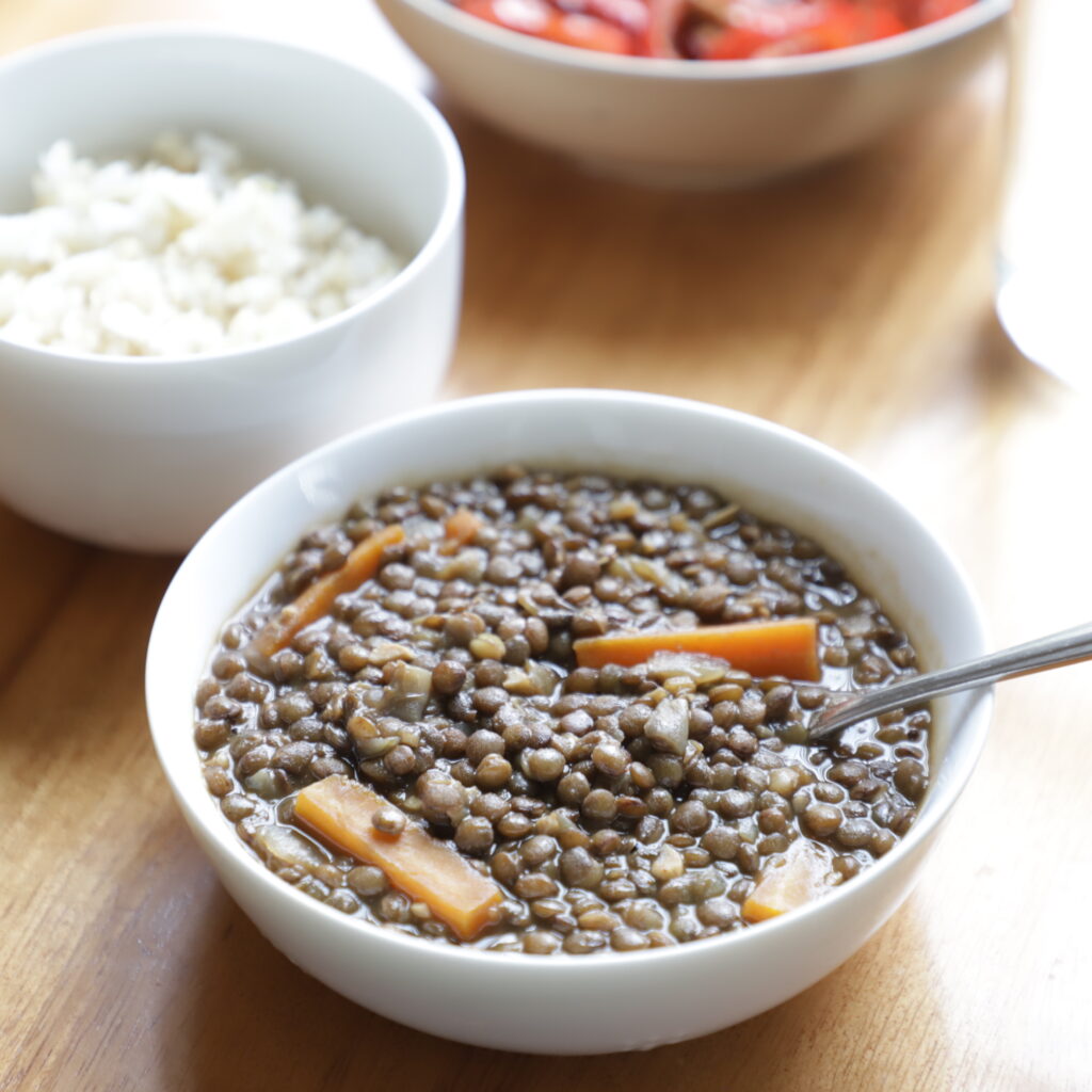 dull-brown small stewed lentils in a low white bowl with chunks of orange carrots and glistening in a soft diffused light, next to a bowl of white rice and sautéed red peppers