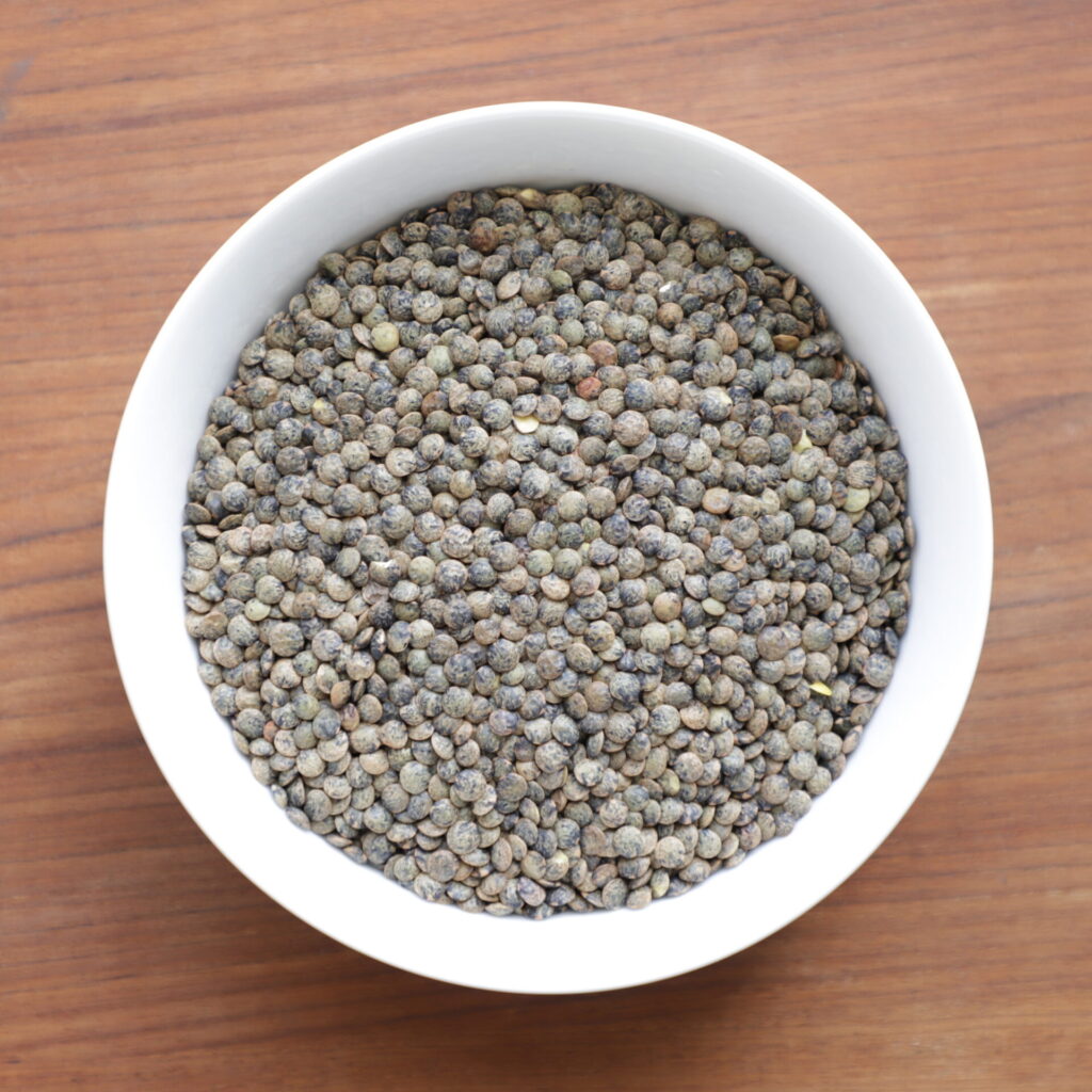 close shot of dull greenish brown French green lentils in a white bowl seen from above on a brown teak background