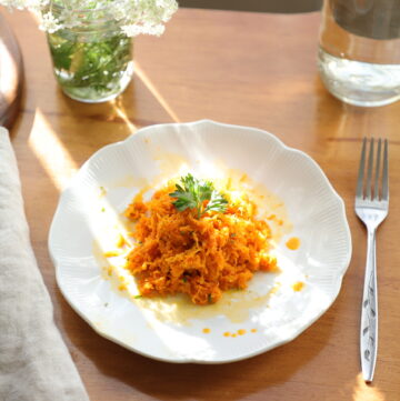 White plate with bright orange carrot salad hit by a ray of sunshine next to a linen cloth, a cup of water, and a vase of wild carrot flowers.