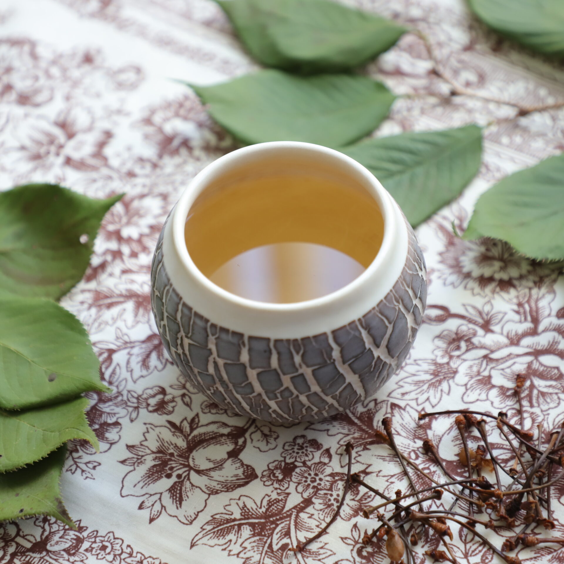 Spherical ceramic cup with white rim and purple crackled effect holding amber liquid next to cherry stems and cherry leaves on a brown flowered tablecloth.