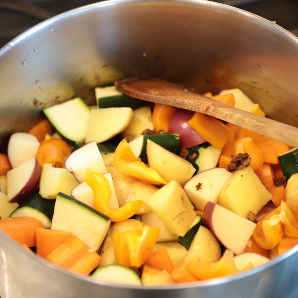 colorful chunks of vegetables in a steel pot, with a wooden spoon 