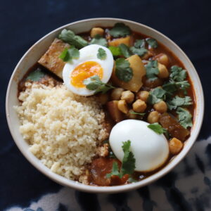 Beige bowl filled with couscous and cooked semolina, and topped with fresh cilantro and two boiled egg halves on a dark blue surface.