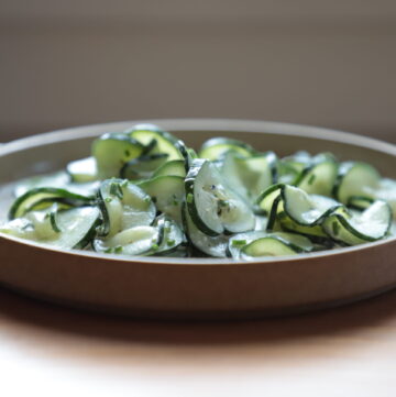 Close up side view of cucumber salad with white dressing and chives on a minimalist brown plate.
