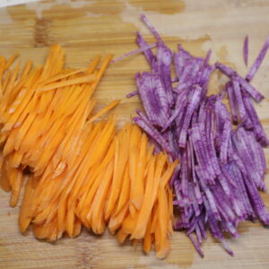 Pile of julienned purple radish and another of carrot on a cutting board.
