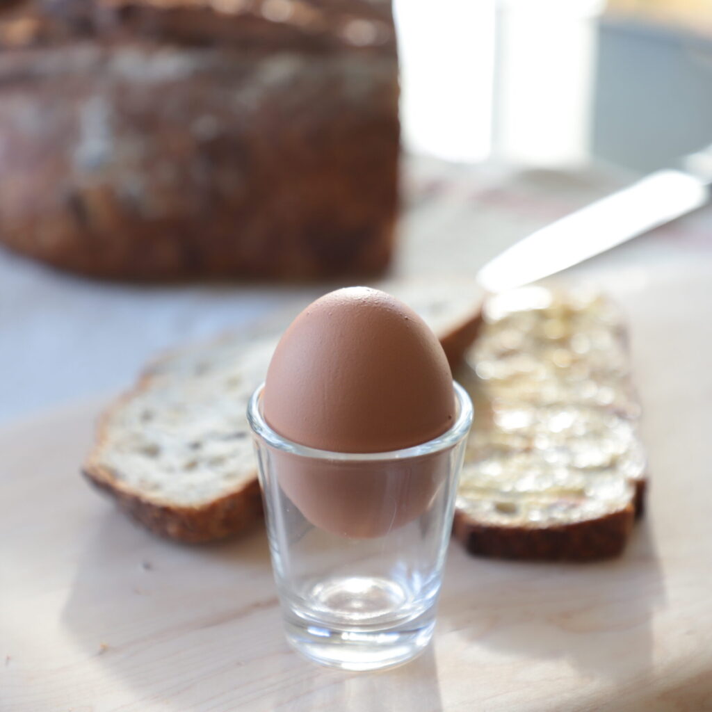 clear shot glass containing soft-boiled egg still whole, 2 pieces of taost behind it, a loaf of bread on a glass table, lit from behind