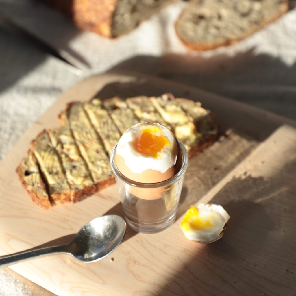 sun-lit soft-boiled egg topped and exposing the orange yolk inside, next to a buttered and sliced toast, a metal spoon, and the top of the egg, on a light colored cutting board and a slice of bread in the background