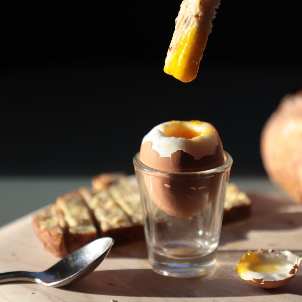 Side view of egg in shell nestled in a clear shot glass, next to a small metal spoon, a piece of buttered bread, and a piece of bread poised in the air exiting the top of the image has just been dipped in the yolk, sunlit from the side and on a black background