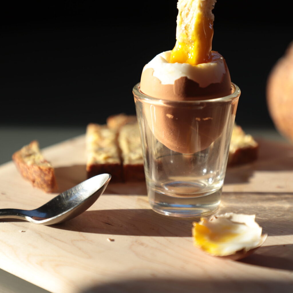 Side view of egg in shell nestled in a clear shot glass, next to a small metal spoon, a piece of buttered bread, and a piece of bread dunked in exiting the top of the image has just been dipped in the yolk, sunlit from the side and on a black background