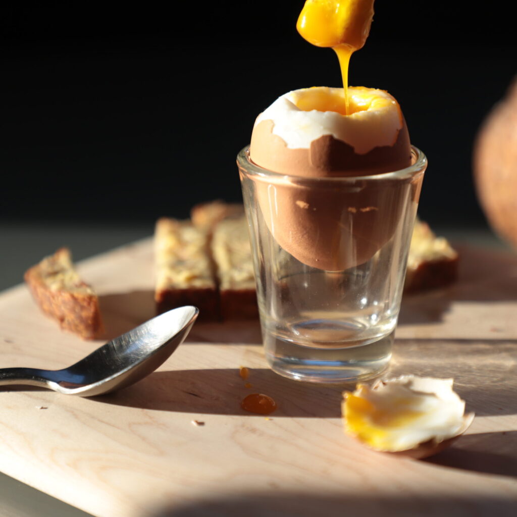 Side view of egg in shell nestled in a clear shot glass, next to a small metal spoon, a piece of buttered bread, and a piece of bread dunked in the yolk and dripping exiting the top of the image has just been dipped in the yolk, sunlit from the side and on a black background