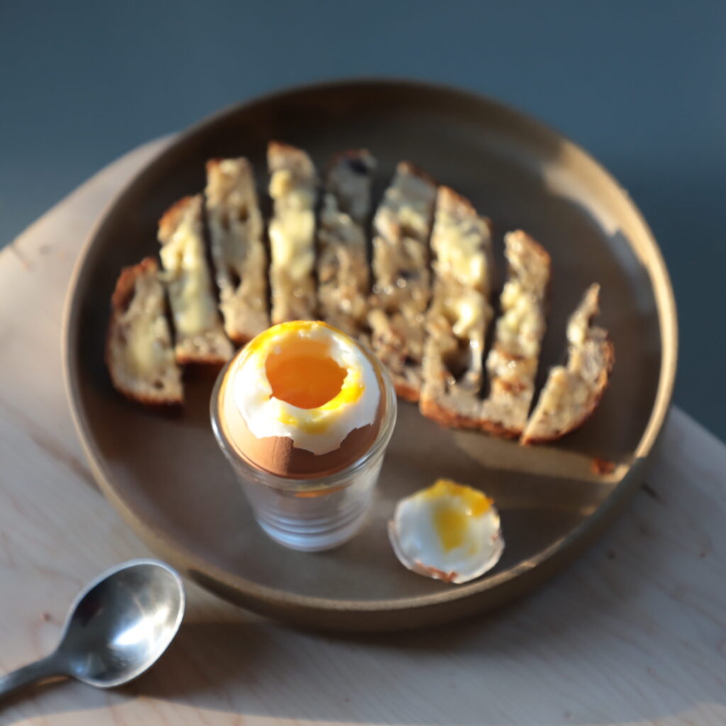 Muted ceramic plate on a pale cutting board, next to a small metal spoon. The plate contains a slice of sourdough buttered and sliced, a shot glass with an egg in shell nested in it, and revealing the inner yolk, and the top of the egg cut off, partly lit by a low, grazing sun