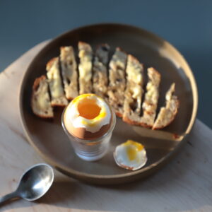 Muted ceramic plate on a pale cutting board, next to a small metal spoon. The plate contains a slice of sourdough buttered and sliced, a shot glass with an egg in shell nested in it, and revealing the inner yolk, and the top of the egg cut off, partly lit by a low, grazing sun.