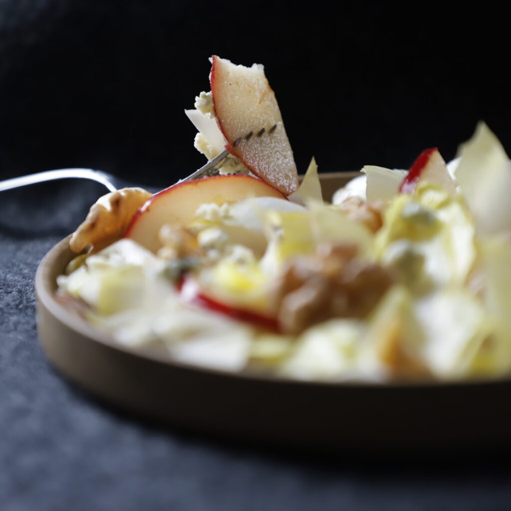 muted brown plate of endive salad with a slice of pear stabbed on a fork and resting on the left side, revealing the moisture on the pear slice