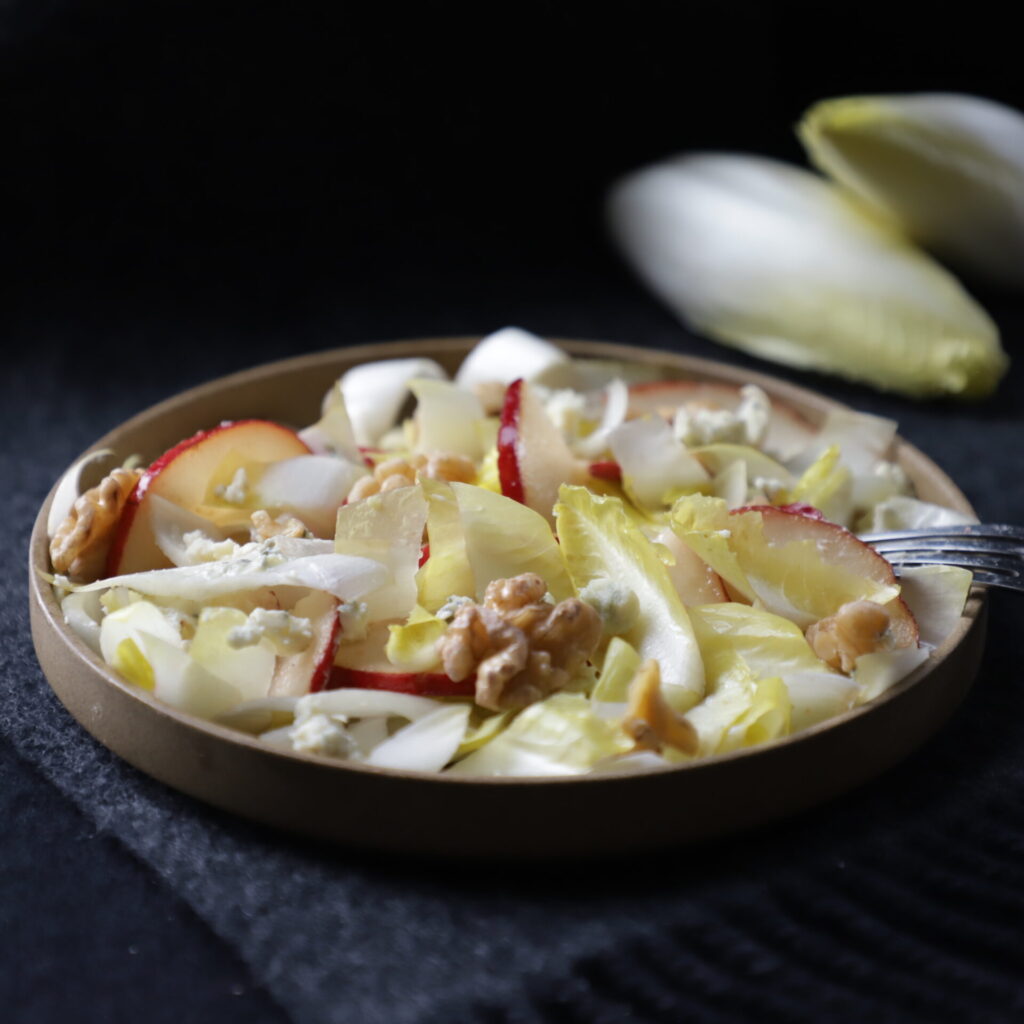 muted brown plate of endive salad on a dark grey cloth and with 2 yellow endives on top of each other in the upper right corner