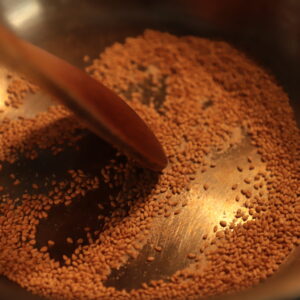 Sesame seeds being stirred with a wooden spoon in a steel skillet, bathed in a warm light.