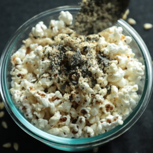 Glass bowl with blue highlights filled with popped corn and being sprinkled with seaweed gomasio.