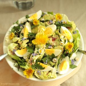 Close up of a white bowl filled with lush dandelion salad topped with boiled eggs pieces and bright dandelion flowers.