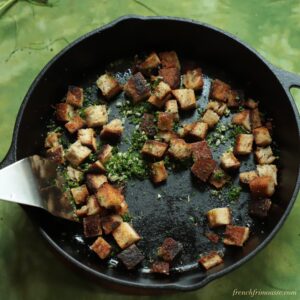 Top view of a black cast iron skillet with herbed croutons and a metal spatula, on a light green background.