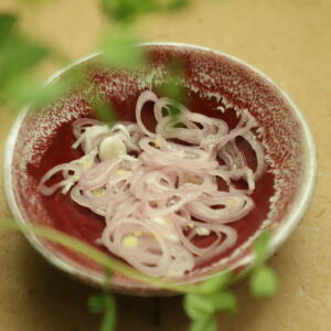 Pickled shallot in a small red bowl with pea shoots blurry in the foreground.
