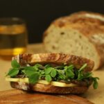 Side view of Portobello Sandwich with pea shoots jutting out, a sourdough loaf and a yellow glass cup in the background.