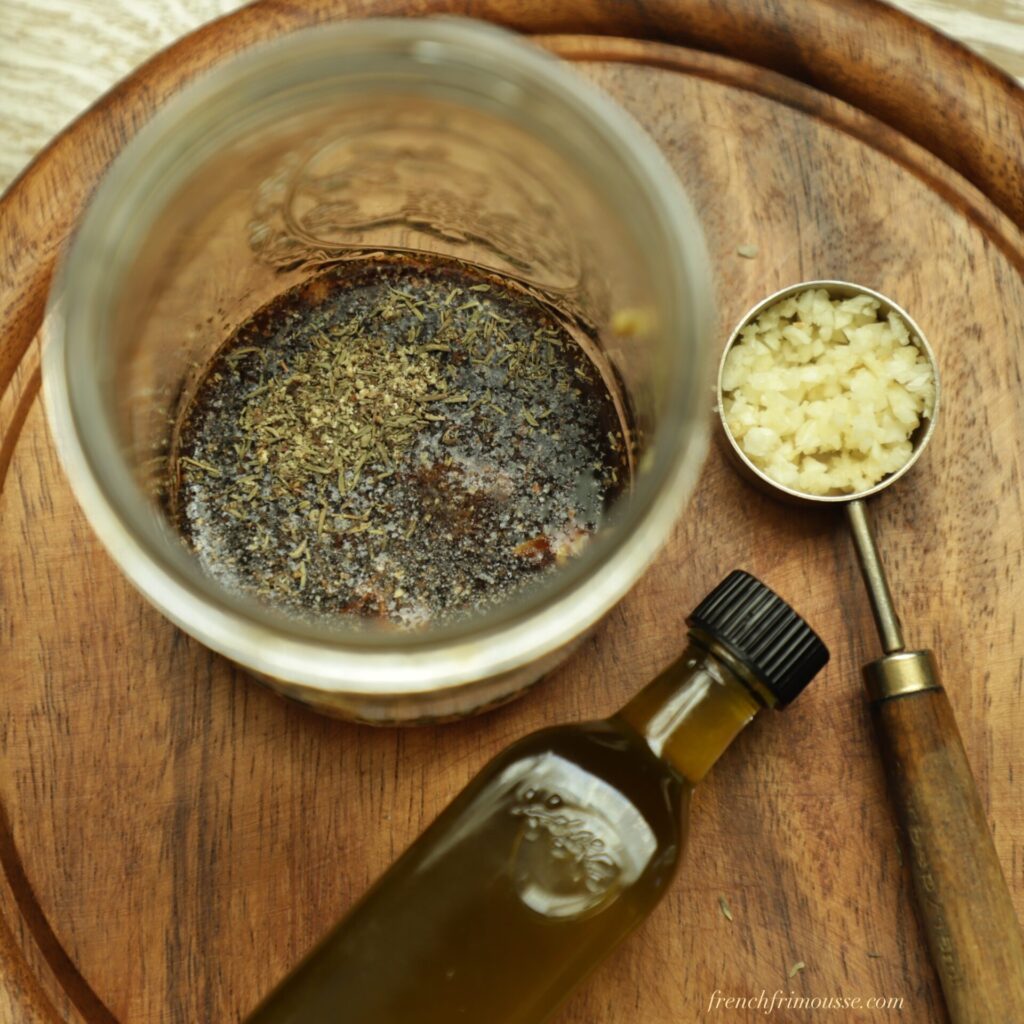 Close up on balsamic dressing in a jar showing thyme leaves and next to a bottle of olive oil and a spoon filled with mined garlic.