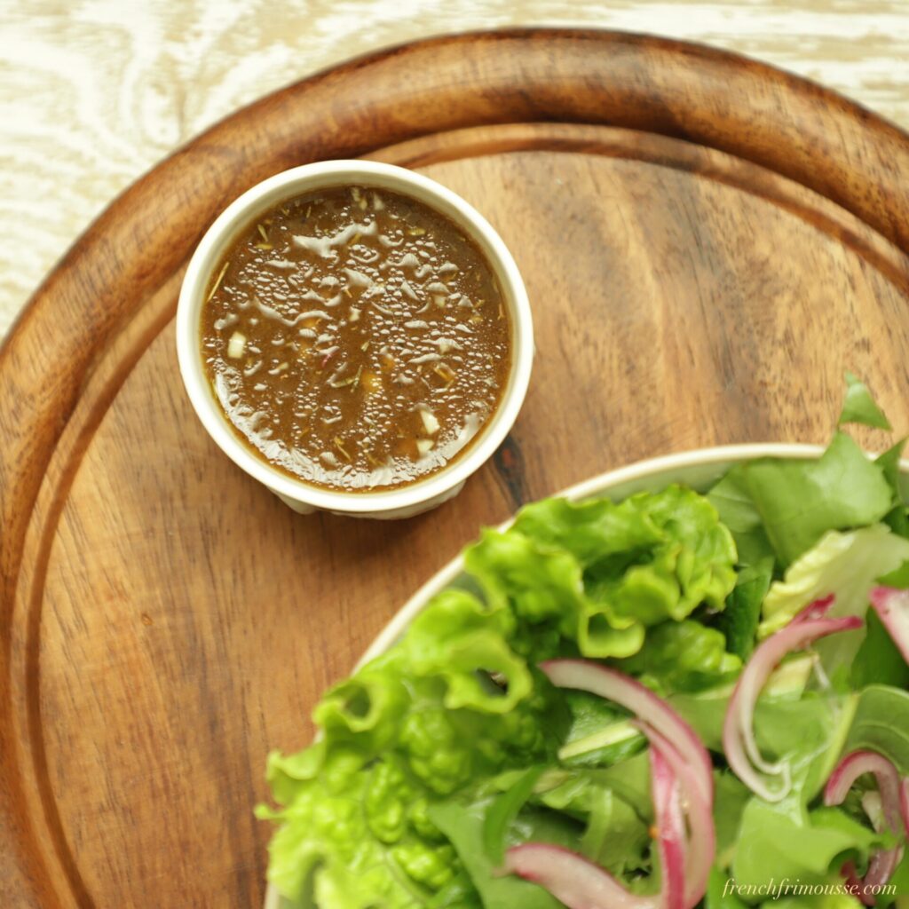 Top view of a round cutting board, a small white bowl of balsamic dressing glistening and part of a bowl filled with bright green salad and sliced red onions.