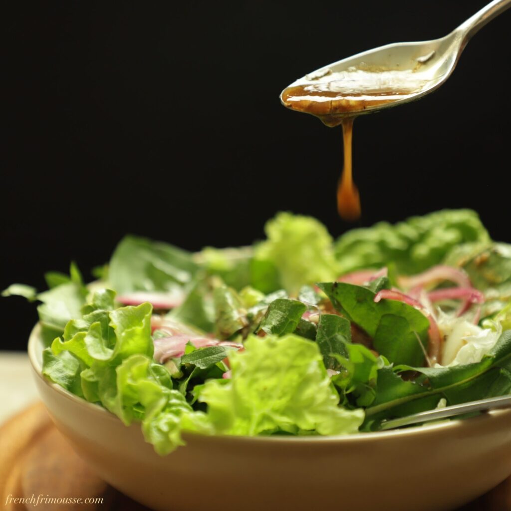 Side view of bright green lettuce and dandelion leaves shone from behind and being drizzled with balsamic dressing from a spoon.