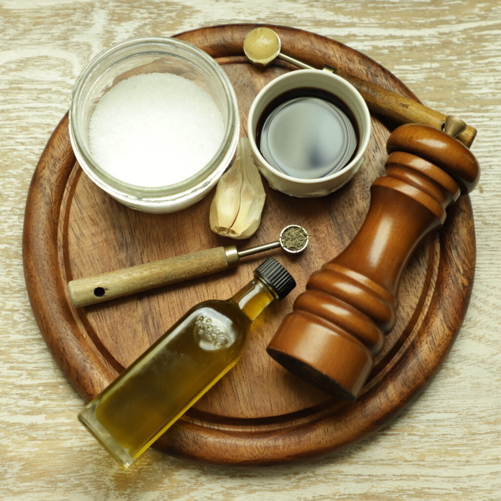 Round cutting board displaying ingredients for balsamic dressing: olive oil bottle, dried thyme, black pepper grinder, salt dish, dark balsamic vinegar in a white dish, mustard, and garlic cloves.